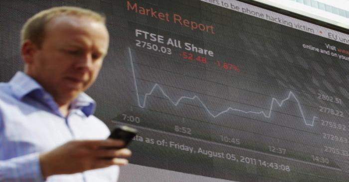 A man passes a screen showing the activity of the FTSE index at Canary Wharf financial district