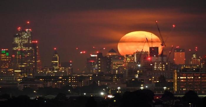 The full moon is seen rising behind skyscrapers at Canary Wharf and the London skyline, London,