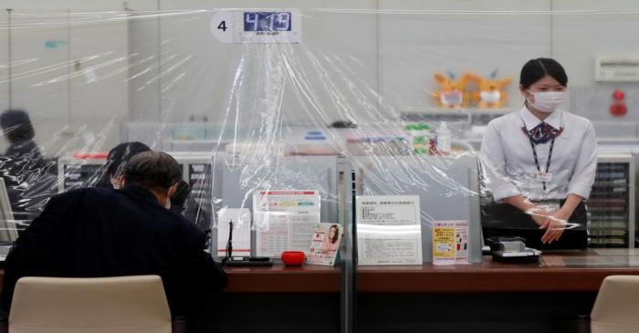 A bank teller wearing a protective face mask stands at a counter where a plastic curtain is