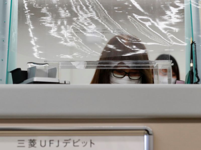 A bank teller wearing a protective face mask works at a counter where a plastic curtain is